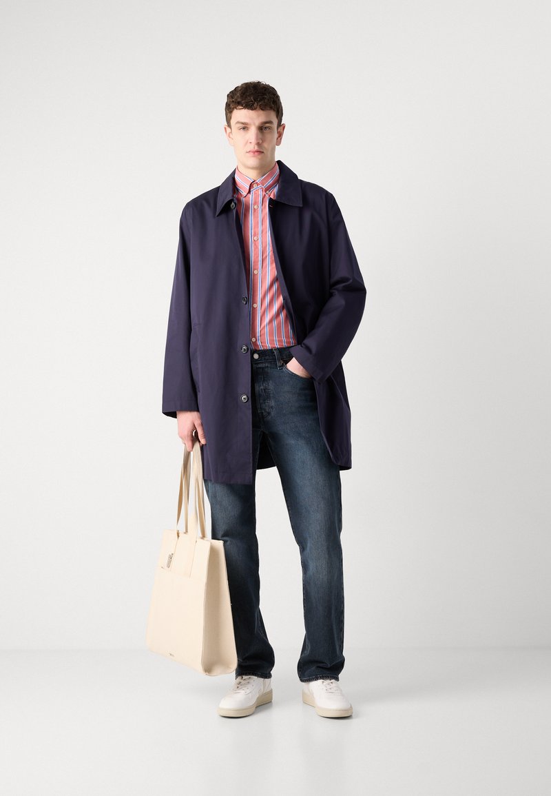 Granatowy płaszcz, koszula w czerwono-białe paski, niebieskie dżinsy, białe trampki, trzymając beżową torbę na ramię, stojąc na jednolitym tle.