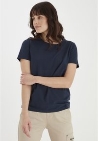 Tricou cu mâneci scurte de culoare bleumarin, confecționat dintr-un material moale, cu guler rotund și croială lejeră, purtat cu pantaloni bej deschis ce au buzunare tip cargo.