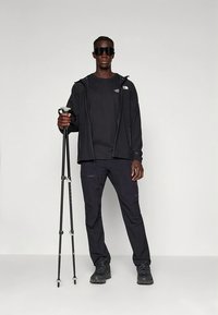 Conjunto de ropa de exterior negra que incluye chaqueta, camiseta y pantalones. Accesorios: gafas de sol negras y bastones de trekking. Textura suave, diseño funcional.