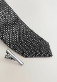 Sort mønstret slips med hexagonale designs, lavet af vævet materiale, parret med en sølv slipsenål med en rektangulær hvid akcent.