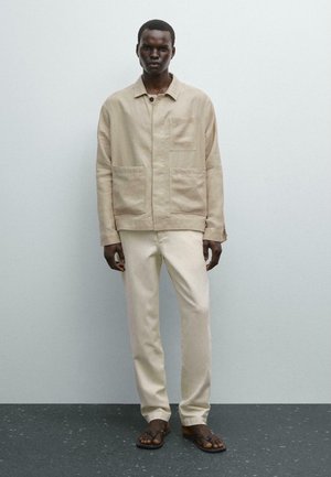 Light beige linen jacket with two front pockets and a buttoned collar, paired with matching beige trousers and brown flip-flops.