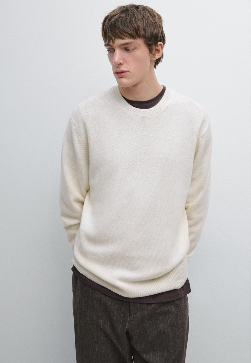 Light beige knit sweater with a round neckline and ribbed cuffs. Worn over a dark shirt, paired with pinstriped dark trousers.