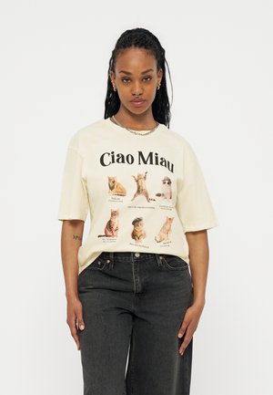 Γυναίκα που φοράει κρεμ μπλουζάκι με το κείμενο «Ciao Miau» και έξι διαφορετικές φωτογραφίες γάτων, συνδυασμένο με φαρδιά μαύρα τζιν.
