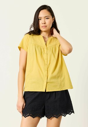 Žena v krátkym rukávom žltom blúzke so zapínaním na gombíky a čiernych šortkách s oblými lemami a detailmi z čipky s dierkami, stojaca pred jednoduchým pozadím.