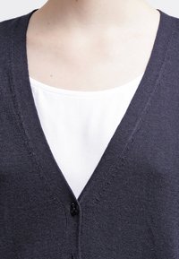 Mörkgrå cardigan med V-hals och knappstängning, över en vit topp. Stickad textur med subtila sömdetaljer.