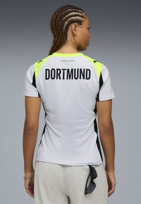 Camisa atlética blanca con acentos en negro y amarillo neón. La parte posterior presenta "DORTMUND" en letras negras y en negrita. Diseñada para actividades deportivas.