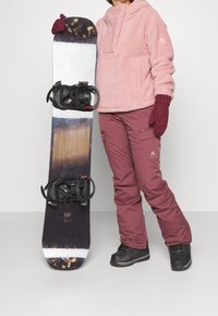 Růžový fleecový pulovr a bordové sněhové kalhoty v kombinaci s černými sněhovými botami. Snowboard má tmavé dřevěné vzory s černými vázáními.