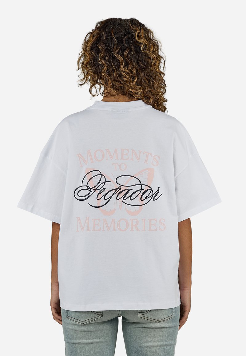 Άτομο με σγουρά μαλλιά που φοράει ένα άνετο λευκό T-shirt με μια πεταλούδα και το κείμενο «Moments to Memories» και «Pegador» στην πλάτη.