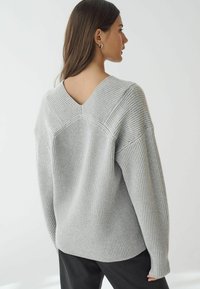 Hellgrauer, gerippter Pullover mit langen Ärmeln, V-förmigem Ausschnitt hinten und strukturiertem Stoff. Lockere Passform und seitliche Schlitze am Saum.