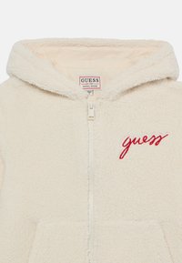 Kremna flis jopica s zip zapiranjem, podložen notranji del in rdeč vezen "guess" logotip na levi strani. Mehka tekstura.
