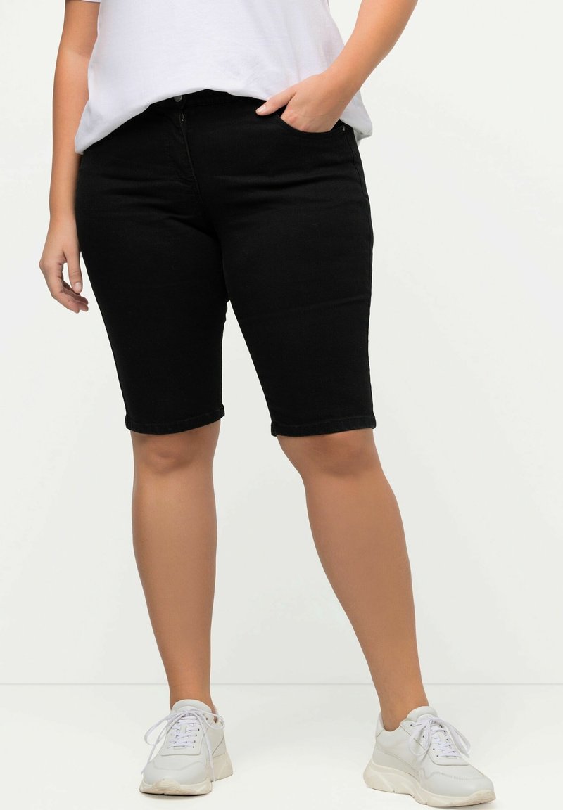 Personne portant un short noir jusqu'aux genoux, un t-shirt blanc et des baskets blanches, debout avec une main dans la poche, sur fond blanc.