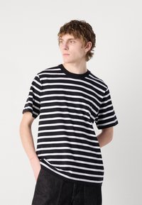 Tricou din bumbac cu dungi alb-negru, cu mâneci scurte și guler rotund; prezintă o croială lejeră și detalii de cusătură vizibile.