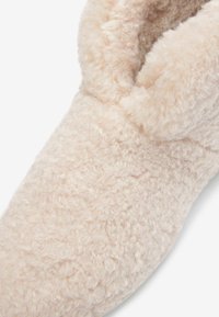 Chausson beige duveteux avec un haut ajusté et une surface texturée, conçu pour le confort avec un matériau doux et moelleux.
