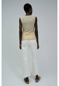 Vrouw met kort haar, gekleed in een beige mouwloos patroon top, witte cropped jeans en beige instap schoenen, staand tegen een effen achtergrond.