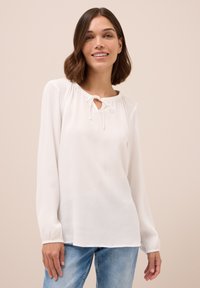 Blusa bianca a maniche lunghe con scollatura rotonda e dettaglio a fiocco, tessuto arricciato sulle spalle e polsini elastici; materiale leggero.