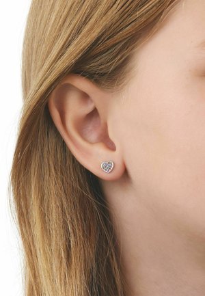 Primer plano de una oreja rubia con un pequeño pendiente en forma de corazón con piedras preciosas multicolores.