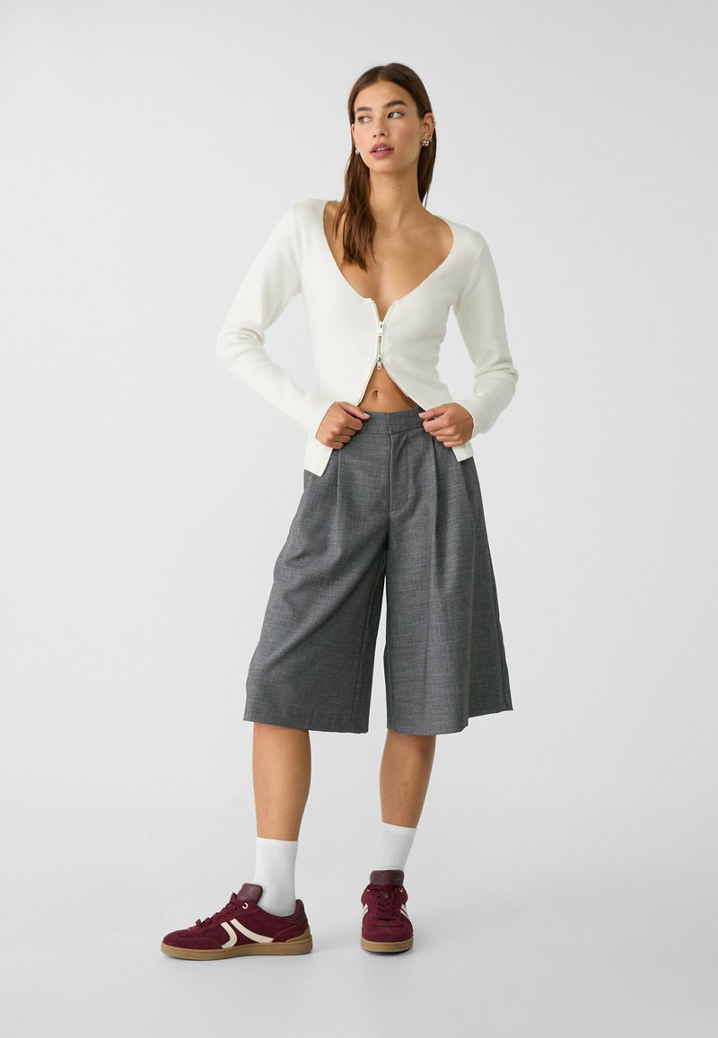 Cardigan bianco con profondo scollo a V e chiusura con zip, abbinato a culotte plissettate grigie e sneakers bordeaux con accenti bianchi.