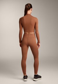 Bruine, aansluitende top met lange mouwen en hoge kraag, gecombineerd met bijpassende leggings. Model draagt zwarte sportschoenen.