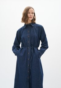 Denim haljina košulja u tamnoplavoj boji. Ima gumbe sprijeda, duge rukave, ovratnik i vezanu struk. Džepovi s bočnih strana.