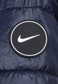 Ναυτικό μπλε μπουφάν με μαλλί, που διαθέτει έναν κυκλικό μαύρο μπατζάκι με το άσπρο λογότυπο της Nike, σε φόντο υφής με γυαλιστερή επιφάνεια.