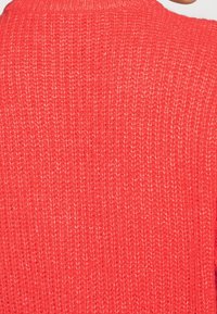 Close-up van een rode gebreide trui met textuur, waarop de gedetailleerde stiksels en het stofpatroon te zien zijn.