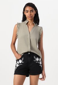 Kaki ermeløs bluse med volangdetaljer på skuldrene; kombinert med svarte shortser med hvit blomsterbroderi på lommene.