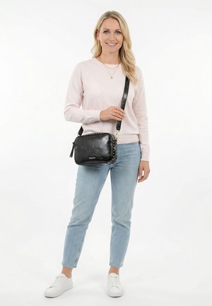Woman wearing light pink sweater, light blue jeans, white sneakers, and a black crossbody bag with studs, standing and smiling on white background.