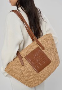 Femme aux longs cheveux foncés portant un pull blanc et un grand sac fourre-tout tressé couleur fauve avec des anses en cuir marron et un patch carré portant l'inscription "LANCASTER"