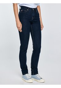 Persona, kas valkā tumši zilas šauras džinsa bikses un gaiši zilas augstās sporta kurpes, stāv pret vienveidīgu gaišu fonu.