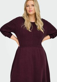 Sarkanvīns kleita ar ribotu tekstūru augšējā daļā, apaļu kakla izgriezumu un garām piedurknēm. Brīvi plūst no jostas.