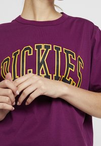 Camiseta de algodón morada con un gran gráfico en negro y amarillo de "DICKIES" en la parte frontal. Manga corta, cuello redondo y textura suave.