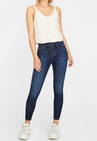 Bílý krajkový crop top s výstřihem do V a detaily knoflíků, spárovaný s tmavě modrými skinny džínami a bílými teniskami. Hladké textury a přiléhavý design.