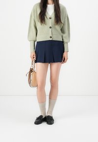 Kobieta w jasnozielonym kardiganie na guziki, granatowej plisowanej mini spódnicy, beżowych skarpetkach, czarnych mokasynach, trzymająca beżową torebkę przy boku.