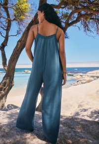 Tuta fluida e ampia in teal smorzato, realizzata in tessuto leggero. Caratterizzata da spalline sottili regolabili e finitura testurizzata.