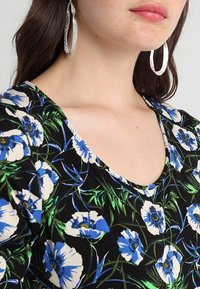 Haut en tissu à motifs floraux présentant des fleurs bleues et blanches sur fond noir, avec un décolleté arrondi. Des boucles d'oreilles en argent en forme de créole sont visibles.