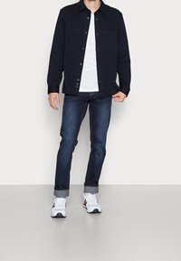 Chemise marine boutonnée sur un t-shirt blanc, associée à un jean en denim foncé et des baskets blanches. Dispose de deux poches avant et de manches retroussées.