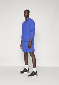 Blauwe sporthoodie en shorts gemaakt van ademend materiaal, gecombineerd met zwarte sneakers met groene accenten en witte sokken.