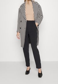 Cappotto in lana bianco e nero a motivo houndstooth con risvoltini ampi, due bottoni e tasche laterali, indossato sopra un top a coste beige e pantaloni neri.