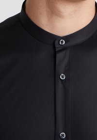 Camicia nera con collo alla coreana, texture liscia e due bottoni con accenti blu. Dettaglio ravvicinato della superficie del tessuto.