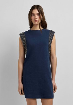 Giovane donna che indossa un vestito senza maniche blu navy con spalline impreziosite, in piedi davanti a uno sfondo semplice di colore grigio chiaro.