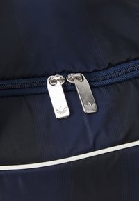 Marineblå nylonveske med teksturerte striper, med doble sølvglidelåser og Adidas-logo samt hvit piping langs sømmen.