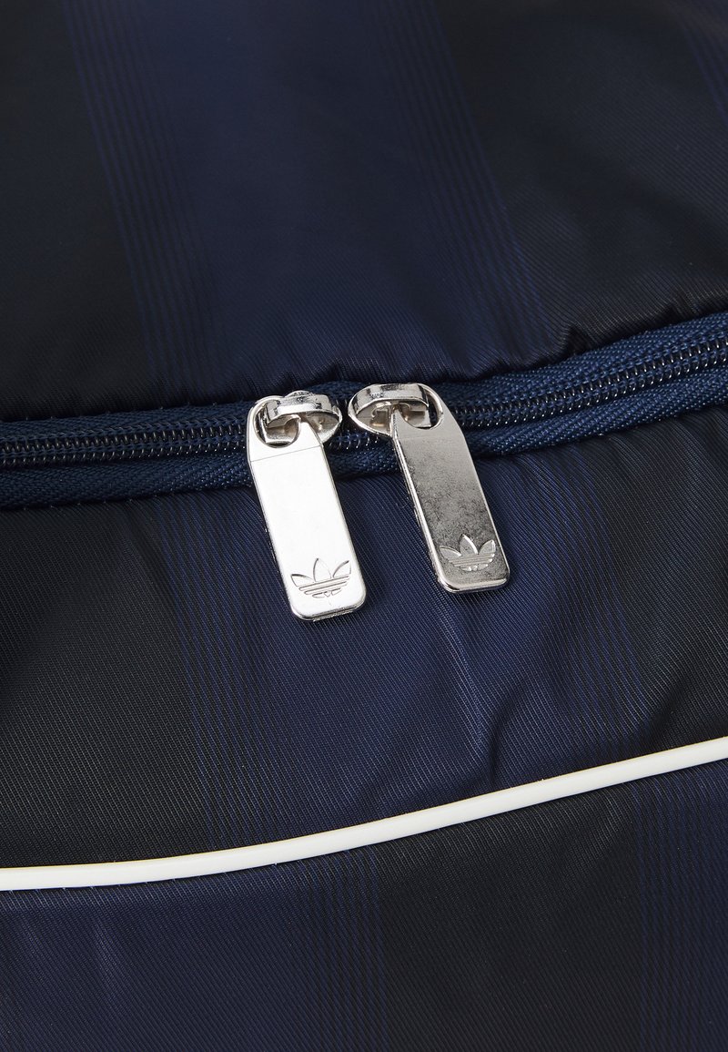 Sac en nylon bleu marine avec des rayures texturées, équipé de deux zips argentés et d'un logo Adidas, avec un passepoil blanc le long de la couture.