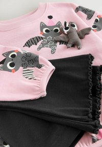 Rosa sweatshirt med tecknade katter och en leksaksbat, kombinerad med svarta byxor med volanger, ordnat snyggt.