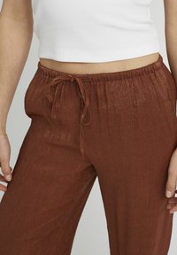 Pantalons légers de couleur rouille, texturés avec une taille élastique et un cordon de serrage à nouer. Présente un ribbing vertical pour le détail.