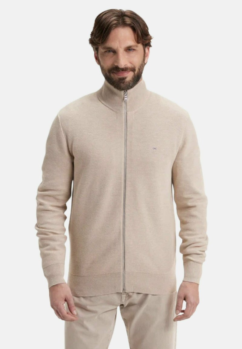 Homme barbu portant un pull beige zippé intégralement et un pantalon beige assorti, debout devant un fond clair uni.
