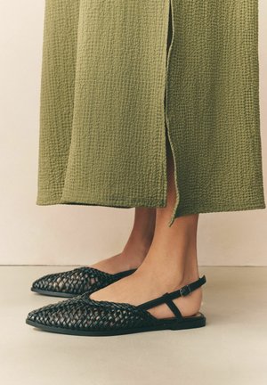 Barfuß mit schwarzen, gewebten Slingback-Ballerinas kombiniert mit einer grünen, strukturierten, weit geschnittenen Hose mit einem vorderen Schlitz.