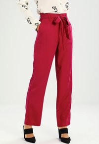 Pantalon taille haute fuchsia avec ceinture nouée devant, coupe cintrée, poches latérales et tissu lisse. Associé à des chaussures noires à talons blocs.