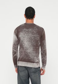 Mann, der mit dem Rücken zur Kamera steht, trägt einen braun-grauen, ausgewaschenen Langarmpullover und dunkelblaue Jeans vor einem einfarbigen weißen Hintergrund.