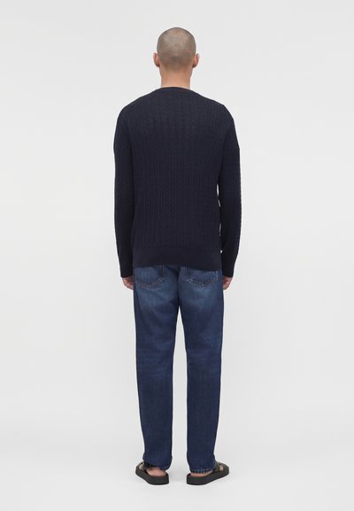 Άνδρας που στέκεται πλάτη, φορώντας ένα σκούρο μπλε πλεκτό πουλόβερ με σχέδιο κοτσίδες, μπλε τζιν και μαύρα σανδάλια σε ένα απλό φωτεινό φόντο.