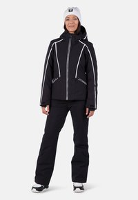 Zwarte geïsoleerde jas met witte accentlijnen, met rits aan de voorkant en capuchon, gecombineerd met zwarte broek en witte beanie, staande op een effen achtergrond.